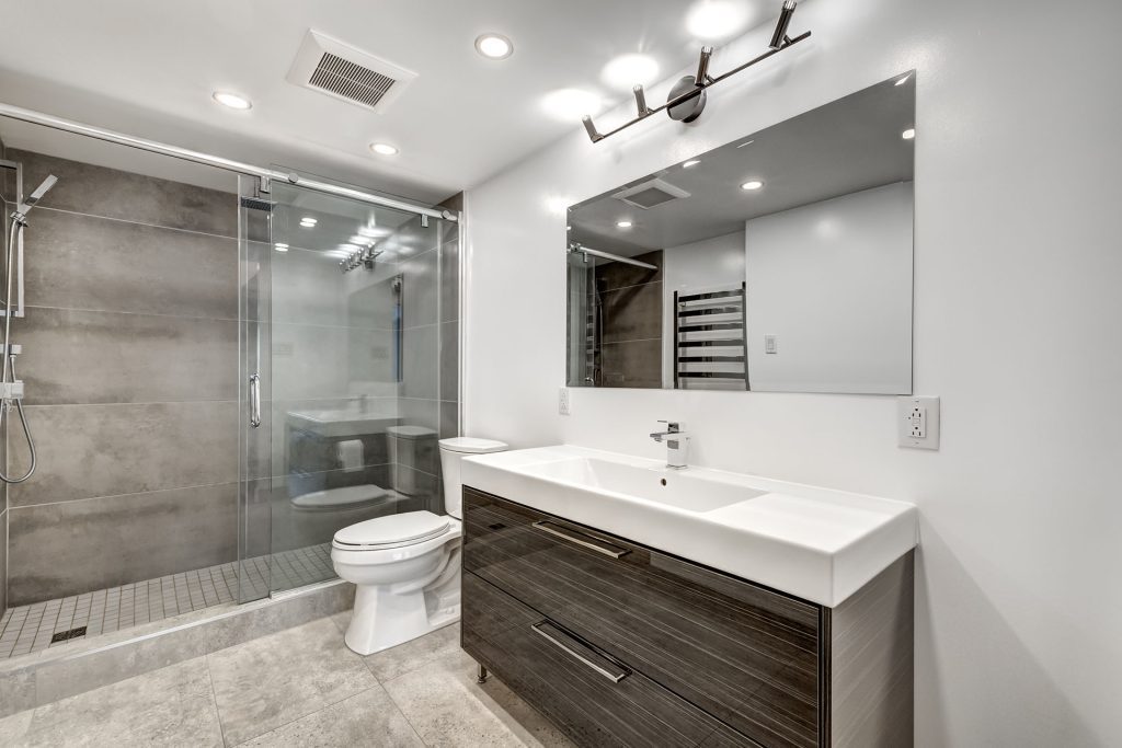 Luxurious bathroom remodel with glass shower door and large mirror and bright lighting with wooden vanity