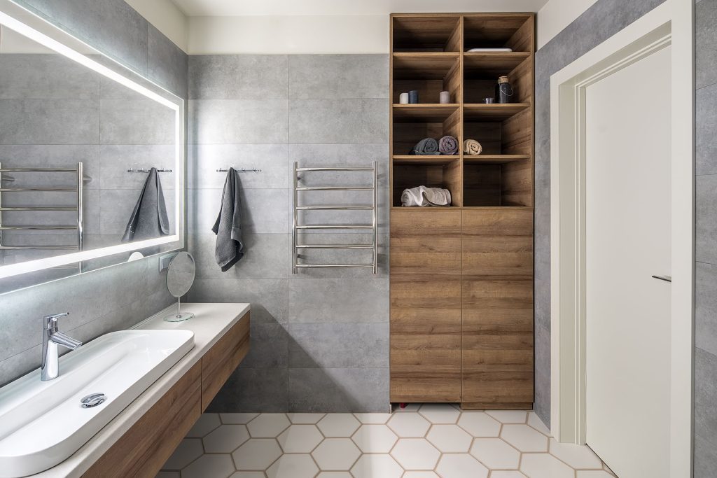 Contemporary bathroom with gray and white tiles. There is large mirror with luminous lamps, tabletop with wooden drawers and sink, towel rack and hanger, wooden locker with shelves with towels, door.
